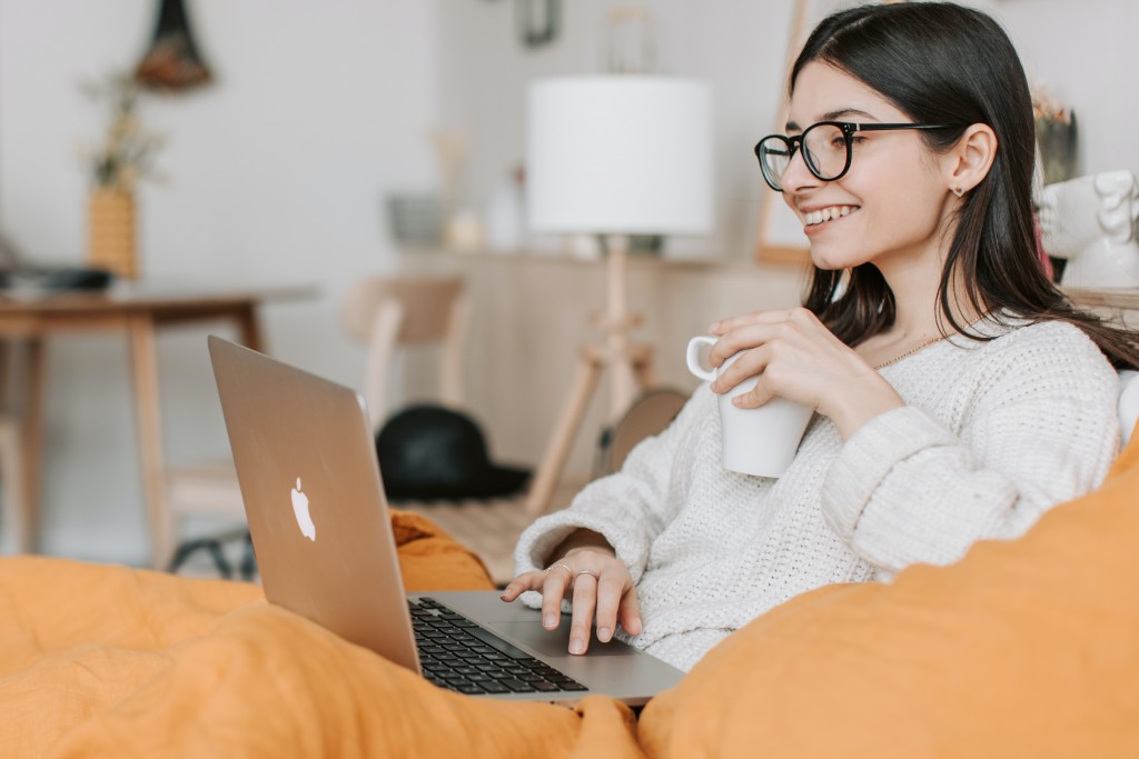 オレンジ色のソファーでコーヒー片手にリラックスしてパソコンを見るメガネをかけた女性